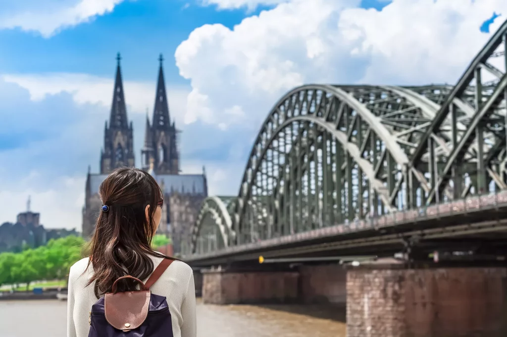 Touristin steht am Rhein und blickt auf die Hohenzollernbrücke und den Dom beim Köln Wochenende