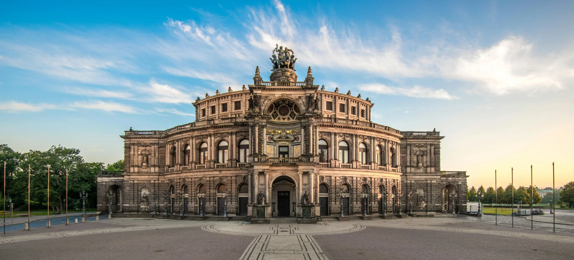 Das Bild zeigt die Semperoper in Dresden, ein Meisterwerk der Architektur und eines der bekanntesten Opernhäuser Deutschlands. Die prachtvolle Fassade des Gebäudes ist von barocker Eleganz geprägt und erstrahlt im warmen Licht des Sonnenuntergangs. Die Semperoper, die auf dem Theaterplatz in der historischen Altstadt von Dresden liegt, wird von einer beeindruckenden Skulpturenreihe flankiert, die den monumentalen Charakter des Gebäudes unterstreicht. Die Architektur spiegelt die Bedeutung dieses Kulturdenkmals wider, das nicht nur für seine Musikaufführungen berühmt ist, sondern auch für seine historische Bedeutung in der Stadt Dresden. Der weite Platz und der klare Himmel im Hintergrund ergänzen das imposante Erscheinungsbild der Oper.